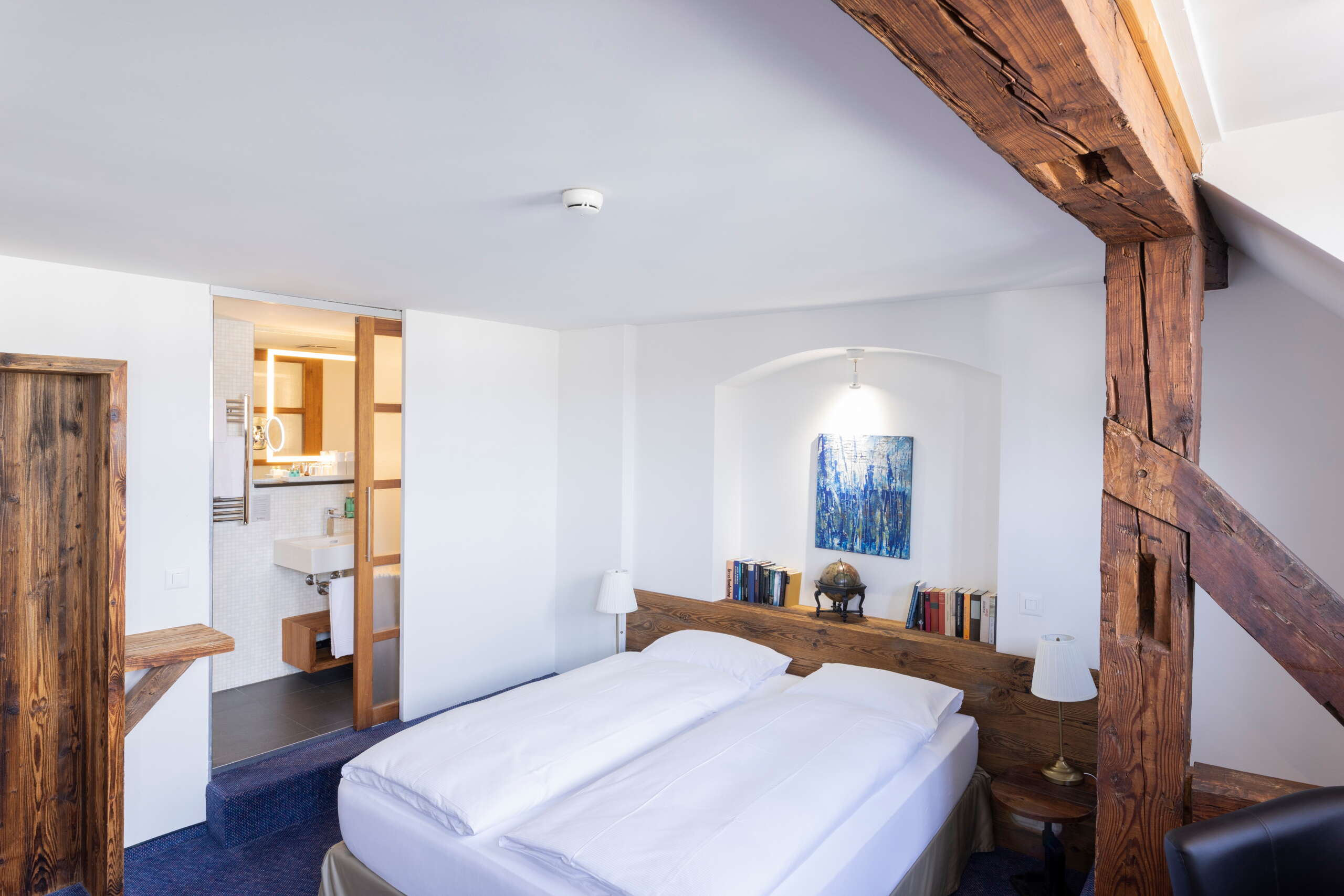 Ein modernes Hotelzimmer mit alten Sichtbalken und einer echten Steinmauer mit einem Doppelbett und einem schmalen Schreibtisch.